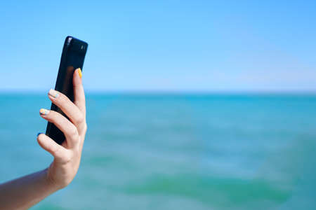 Mobile phone in womans hand. Girl holds smartphone on sea background. Sunny summer day. Roaming and...