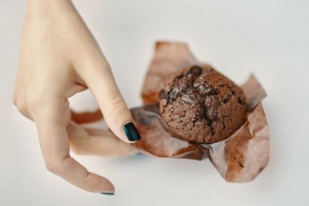Womans hand picks up chocolate cupcake from table. Dark chocolate dessert in paper wrapper. Breakfast, sweet food concept. Sweet muffin on white background.の写真素材