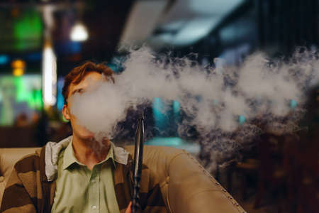 Young guy in cafe smoking hookah. Thick smoke from shisha. Man on sofa in restaurant. Neon signs on...
