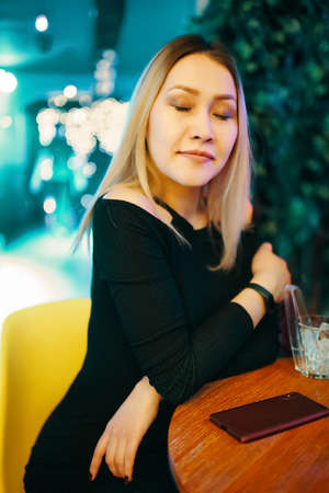 Woman with closed eyes is sitting at table in lounge bar with mobile phone. Beautiful girl in black dress in restaurant. Blurry lights on background. Party and leisure time at cafe.の写真素材