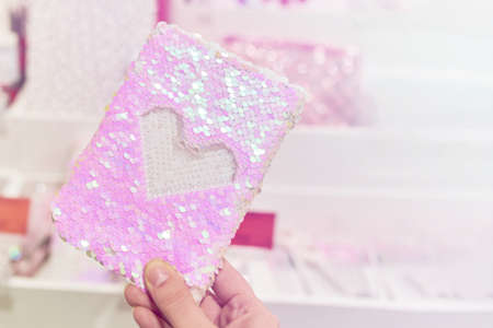Notebook with anti-stress sequin cover in woman hand. White heart on pink artwork. Selling cute...