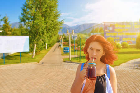 Red-haired woman in dress drinks cocktail on beach. Alley with trees and green lawn. Beautiful girl...