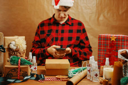 Man in plaid shirt and Santa hat with mobile phone in holiday decorations. Table with gift boxes, antiseptics, medical masks and sweets. New years preparations. Christmas in quarantine.の写真素材