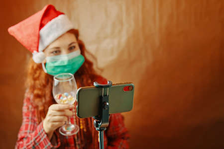 Woman in medical mask and Santa Claus hat with glass of pills in front of her smartphone. Christmas video calls are quarantined. New years mood.の写真素材