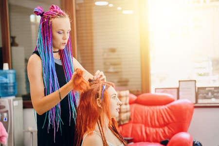 Barber braids dreadlocks. Hairdresser weaves to redhead female ginger afro pigtails and speak with her. Beauty salon services. Hippie and boho style coiffure with kanekalon.の写真素材