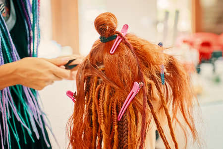 Barber braids dreadlocks. The hairdresser weaves braids with red kanekalon beautiful red-head girl. Beautiful cool hairstyles. Hair service in a beauty salon. Fashionable womens style.の写真素材