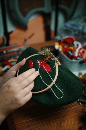 Female hands doing stitch. Process of embroidery of mushroom hat in wooden hoop on green material. Concept of craft diy, hobby, leisure. Accessories for needlework on background.の写真素材