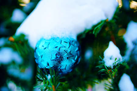 Blue shiny ball with stars on Christmas tree covered in snow. Winter festival atmosphere. Street holiday decorations. New Years aesthetic background for postcards.の写真素材