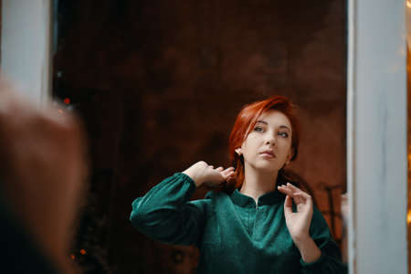 Noble red-haired girl admires herself in mirror. Fabulous retro girl in velvet green dress sits and adjusts her coiffure. Vintage brown wall on background.の写真素材