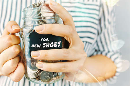 Female in striped shirt with jar of coins in hands. Glass bottle with black sticker. Girl saves money for new shoes. Womens whims. Savings to meet household needs.の写真素材