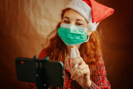 Red-haired woman in Santa hat and medical mask sprays antiseptic on her smartphone. Christmas in quarantine. Video calls via mobile phone.の写真素材
