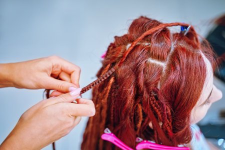 Beauty salon services. Girl hairdresser weaves woman ginger dreadlocks. Braiding process plait with...