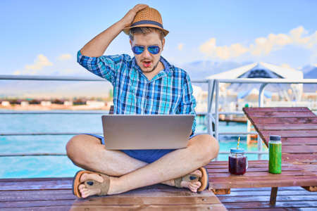 Concept work on vacation. Man is sitting on the pier and looking at the laptop computer. Freelancer in the summer at the sea. Bottle of detox cocktail on the table. Emotions, surprise, delight.の写真素材