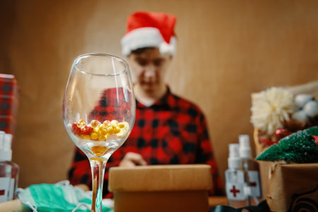 Glass with vitamins in close-up and man in Santa hat in background. Gift boxes, antiseptics and medical masks on table. Guy among Christmas items. New year on quarantine.の写真素材