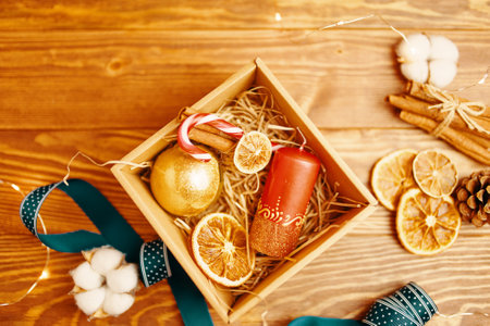 Flat lay of Christmas gift box and decorations on wooden table. New Years atmosphere. Candy cane, candle with ornate, ribbon, dried orange, golden ball, cinnamon and pine cone. Festive still life.の写真素材