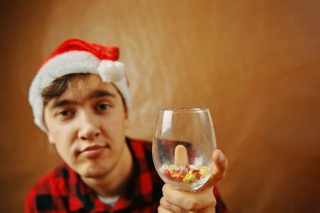 Man in Santa Claus hat holds a glass with pills and drugs. Fighting the coronavirus on holiday. Holidays spending under quarantine. Guy in shirt with pensive grimace on face. New year mood.の写真素材