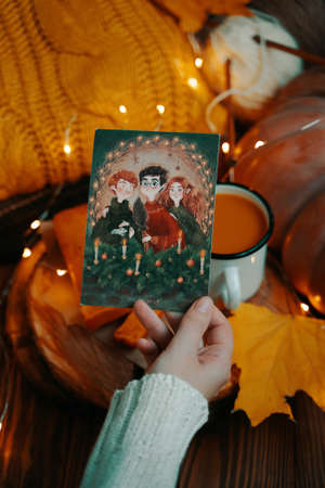 Womans hand in a white blouse holds a card with a Golden trio. Postcard with Harry Potter. Sweater, pumpkin, garland and autumn leaves on wooden background. Bishkek, Kyrgyzstan-November 1, 2019.のeditorial素材