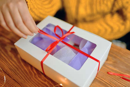 Girl in orange knitted sweater opens festive gift. Christmas or Birthday. Close-up of hand pulling ribbon from box on wooden table.の写真素材