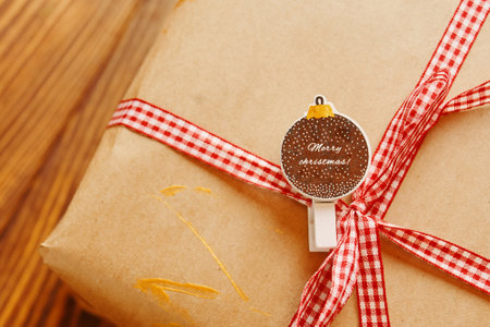 Close-up of gift box with red ribbon and badge on wooden table. Pin with Christmas inscription. Gold patterns on wrapping paper. Festive composition for postcards. New Year mood.の写真素材