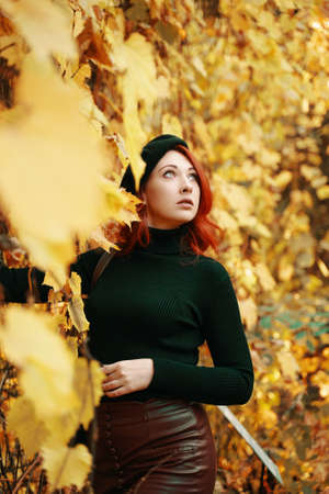 Beautiful young red-haired woman in autumn park. Stylish model among yellow dry foliage of trees. Awesome female in black beret, knitted sweater looks up on sky. Walk in fall nature.の写真素材
