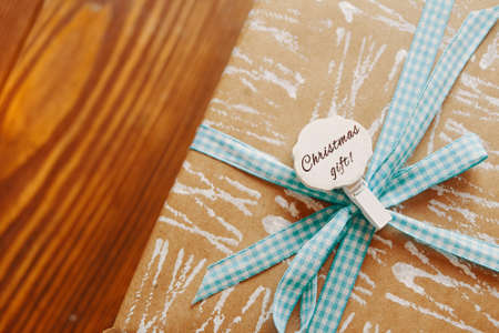Close-up of gift box with ribbon and badge on wooden table. Pin with Christmas inscription. White patterns on wrapping paper. Festive composition for postcards.の写真素材