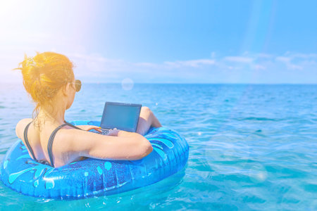 Young red-haired woman is floating on sea in swimming circle. Girl is resting on sea on inflatable ring with mini-computer. Freelancer at sea. The benefits of remote work. Black one-piece swimsuit.の写真素材