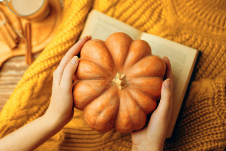 Round ripe pumpkin in hands, hot latte, open romantic book and warm knitted sweater. Autumn composition, top view. The concept of thanksgiving and Halloween. Fall still life, vintage style.の写真素材