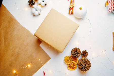 Flat lay of christmas gift box, craft paper and decorations on white textured table. Candy cane, garlands, dried orange slices, cotton and cones. Festive winter still life. New Years atmosphere.の写真素材