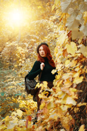 Stylish red-haired female with brown backpack in autumn park. Women in sweater and with beret on head looks away. Bushes and trees with yellow fall foliage. Relaxation, enjoying, solitude with nature.の写真素材
