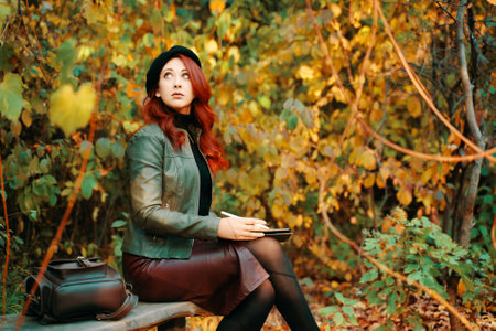 Cute woman with graphic tablet on park bench. Autumn portrait. Red-haired girl in beret and fashionable jacket looks up. Designer or freelancer. Leather backpack on wooden bench.の写真素材