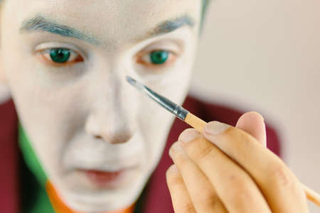 Mim is preparing makeup for performance. Close up of clown cosplayer. Guy with green hair in burgundy suit. Theatre atmosphere. Male actor is covering his face by white greasepaint using brush.の写真素材