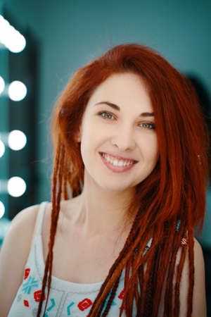 Beautiful woman smiles and looks at camera. Portrait of red-haired girl with dreadlocks in beauty salon. Unusual coiffure. Hippie or boho style. Mirror with light bulbs in background.の写真素材