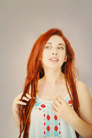 Awesome red-haired girl touching her dreadlocks with fingers. Boho or hippie hairstyle. Woman with pigtails from kanekalon on grey wall background.の写真素材