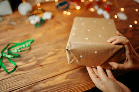 Close up of womens hands packing Christmas gift on wooden table. Wrapping craft paper with white...