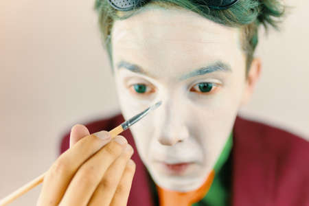 Mim is preparing makeup for performance. Clown cosplayer concept. Man with green hair in burgundy suit. Theatre atmosphere. Male actor is covering his face by white greasepaint using brush.の写真素材