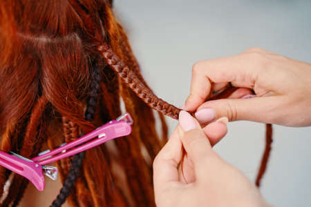 Hairdresser weaves woman ginger dreadlocks. Hippie style hairstyle. Bright pigtails on head. Beauty salon services.の写真素材