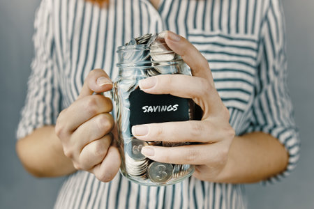 Girl n white and black striped shirt holds her savings. Glass bottle with silver coins in womens hands. Sticker with inscription on money jar.の写真素材
