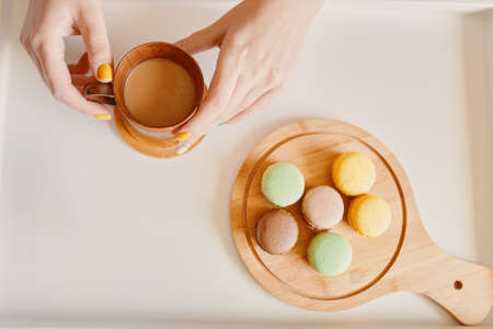 Morning breakfast on white table. Cup of coffee with milk in woman's hands and wooden tray with macaroons. Sweet dessert and hot drink.の写真素材