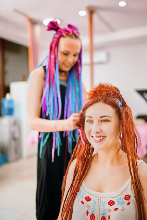 Woman hairdresser with colored afro braids weaves to funny redhead girl ginger dreadlocks. Beauty salon services. Hippie and boho style coiffure with kanekalon.の写真素材