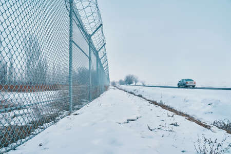 Private secured object near highway. Maximum security detention facility. Car goes on road. Barbed wire fence on border in winter. Closing for quarantine. Unauthorized entry is prohibited.の写真素材