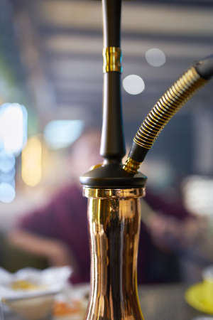 Hookah on table in lounge bar. Close-up of black pipe and golden shisha flask. Smoking person in blurry background. Leisure in oriental cafe.の写真素材
