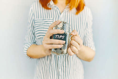 Glass bottle with silver coins in womens hands. Sticker with inscription on money jar. Redhead girl n white and black striped shirt holds her tips pot.の写真素材