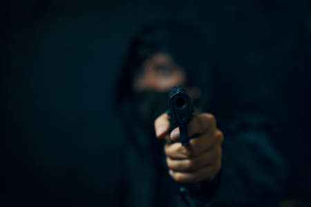 Masked robber with revolver aiming into the camera. Man in hood threatens with firearm. Gun in persons hands. Murderer or armed thief. Criminal with pistol.の写真素材