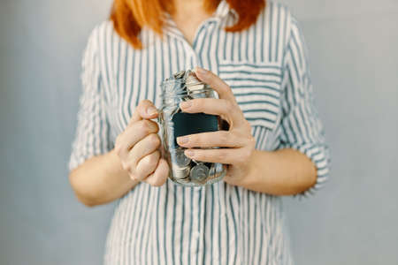 Glass bottle with silver coins in womens hands. Blank black sticker on donation money jar. Redhead girl n white and black striped shirt holds her savings. Pot for tips.の写真素材