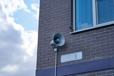 Megaphone hanging on apartment building. City hazard warning system. Loud speaker on brick wall with window. Providing security in town, notification of emergencies. Cloudy sky on background.の写真素材