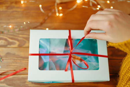 First-person view of womans hand in knitted sweater opening Christmas surprise. Girl pulls ribbon on gift box. Wooden table with garland. New Years holiday mood.の写真素材
