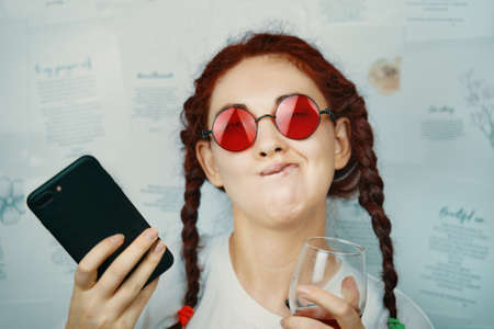 Funny girl with pigtails and red sunglasses makes faces. Female blogger with a glass of wine and a smartphone in her hand. Pampering and antics. Hippie or boho style fashion blog.の写真素材