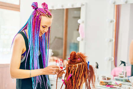 Hippie style hair. Woman hairdresser with colored afro braids weaves to girl ginger dreadlocks. Stylish therapy professional care concept. Weaving process with kanekalon. Beauty salon services.の写真素材