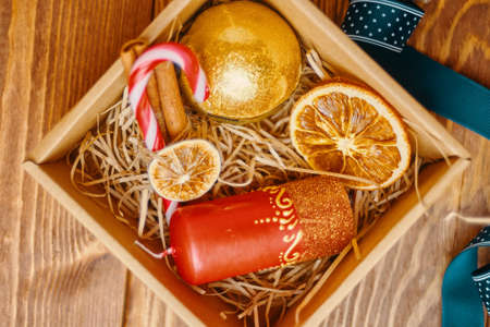 Top view of christmas gift box and decorations on wooden table. New Years atmosphere. Candy cane, candle with ornate, ribbon, dried orange, golden ball, cinnamon. Festive still life.の写真素材