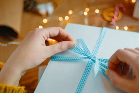 Womens hands tie blue ribbon bow on white gift box. Surprise for Birthday or Christmas. New Years mood. Garland lights and decorations in background.の写真素材
